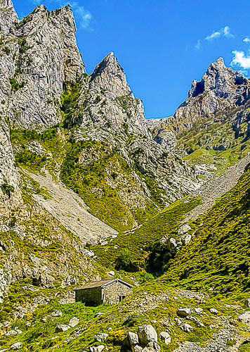 Podemos ver un poco más atrás del primer plano una pequeña casa de piedra (seguramente un pequeño refugio), en plano de fondo podemos ver 3 impresionantes agujas de de granito en la parte izquierda y otra en la parte derecha, con la parte baja cubierta de verde hierba, el cielo que se ve tras ellas es de un color azul intenso.