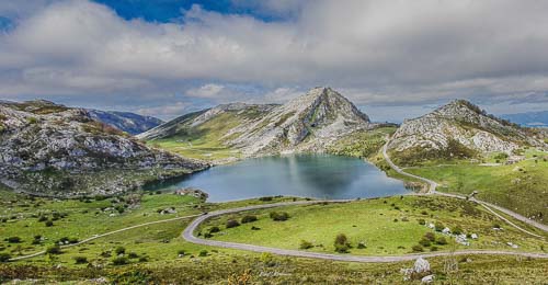 En esta foto se ve en el plano medio y en el centro el lago Enol, en primer plano y hasta la mitad de la imagen se ve hierba y el serpenteante camino que baja hacia el lago, a la izquierda del lago también en el plano medio se ve una pequeña cima de roca gris sin vegetación, en el plano de fondo de mitad a la derecha otras dos cimas igualmente grises y de fondo el cielo azul con unas nubes blancas.