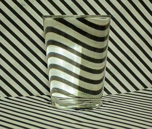 Esta es una simple imagen de una ilusión óptica. En primer plano aparece un vaso lleno hasta arriba de agua que tiene detrás un folio pegado en la pared con rayas diagonales. Por uno de los distintos efectos ópticos de la luz las líneas que quedan en el espacio de dentro del vaso, se ven en vez de en diagonal en horizontal.