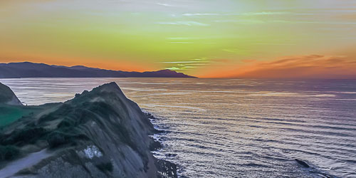 En esta foto del atardecer, se puede ver en primer plano a la izquierda una alargada roca, que llega hasta la mitad de la imagen, en ambos sentidos, en el lado derecho de la imagen y hasta el último plano está el mar azul que según se aproxima al horizonte se vuelve anaranjado. En el lado izquierdo en el plano fina y como horizonte hasta la mitad de la imagen (la otra mitad del horizonte es mar) la costa, pero ya de Bizkaia detrás de la cual ya se ha escondido el sol que tiñe la parte baja del cielo de color naranja.