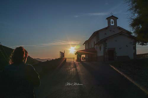 Una imagen obtenida a contraluz. Podemos ver en primer plano y a la izquierda la silueta de una chica de espaldas con una coleta, a la derecha en plano medio la ermita de San Telmo en Zumaia,  en plano de fondo, el sol a media altura, con el denominado en fotografía "efecto estrella", en este caso de 6 puntas.
