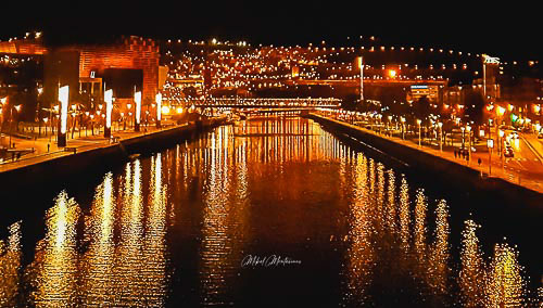 En esta imagen de una apacible noche en Bilbao podemos ver centrada y con las orillas en diagonal desde las esquinas inferiores hacia el centro a media altura de la imagen, una imagen de la ría, en las orillas de la ría y a ambos lados podemos apreciar hileras de farolas con la luz en color naranja y los reflejos de esta luz en el agua de la propia ría.
