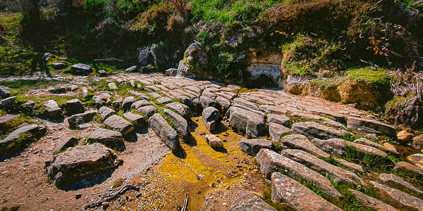 En esta foto se ve en diagonal de la parte superior izquierda a la inferior derecha y hacia nosotros, un enladrillado suelo, pero este es natural con piedras marrones, en la parte inferior izquierda hay un triángulo de arena y por el que corre el agua. En el plano medio y centrada en la foto esta la fuente de a que brota la fresca agua. Por encima de ella y en la misma diagonal de izquierda a derecha tenemos vegetación.