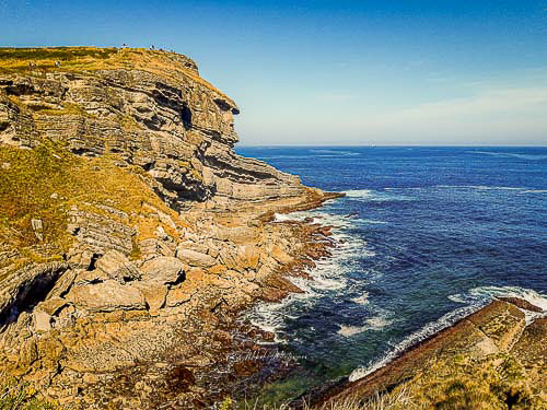 En esta imagen podemos ver en primer plano a la izquierda un acantilado de unos 30 metros, otro de la misma altura, en el plano medio pero a la derecha y en plano de fondo la línea del horizonte del mar, con gran contraste de colores.