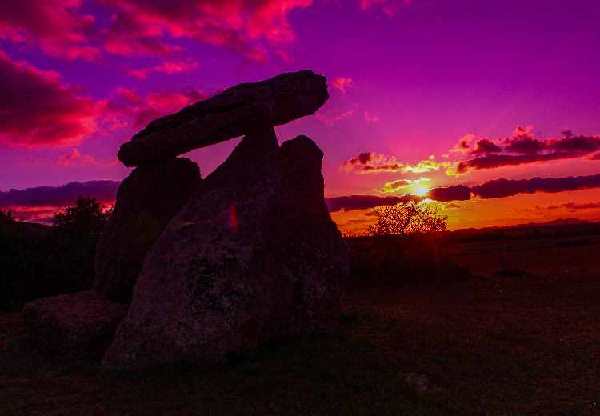 En esta foto de un atardecer en Sorginetxe tenemos en el plano medio el dolmen, en el centro un poco a la izquierda. Detrás está metiéndose el sol a la derecha del dolmen y a la misma altura, pero en el centro de la imagen, en la izquierda en la parte de arriba hay nubes rosadas. Todo el atardecer tiene tonos lilas y rosa.