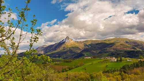En esta foto a la izquierda en primer plano se ven las ramas de un árbol con hojas verdes, en el plano medio y en el centro la cima del puntiagudo monte Txindoki, y hacia la derecha unas lomas tímidamente nevadas, de fondo tenemos un cielo azul con nubes blancas y claros.