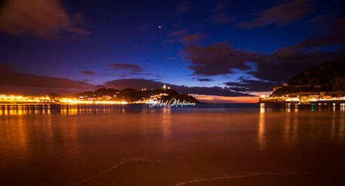En esta foto y hasta la mitad de ella tenemos la arena de la playa con una pequeña ola en el lado derecho y en la parte inferior. En el plano medio a la derecha tenemos la zona del puerto de Donostia, iluminado por la luz de las farolas que se reflejan en el mar. También en el plano medio y en el centro de la imagen se ve la isla de Santa Clara y detrás de ella un poco más a la izquierda el monte Igueldo, a media altura de la foto y hasta la mitad de ella se ve las luces que iluminan el paseo de la Concha, unas luces que se reflejan en el mar. El horizonte divide la foto en dos por encima tiene un cielo de azul intenso mientras que la parte inferior es de color naranja aún.
